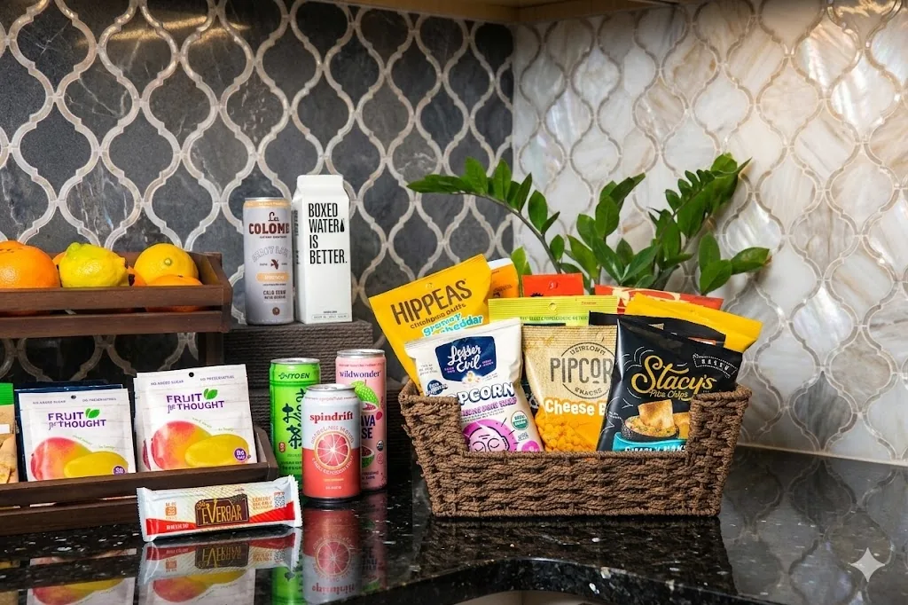Curated office snack arrangement on a dark countertop: a woven basket of healthier brands (Hippeas, Pipcorn, Stacy's, Spindrift, Wildwonder, La Colombe cold brew) next to a wooden crate of fresh oranges and lemons.