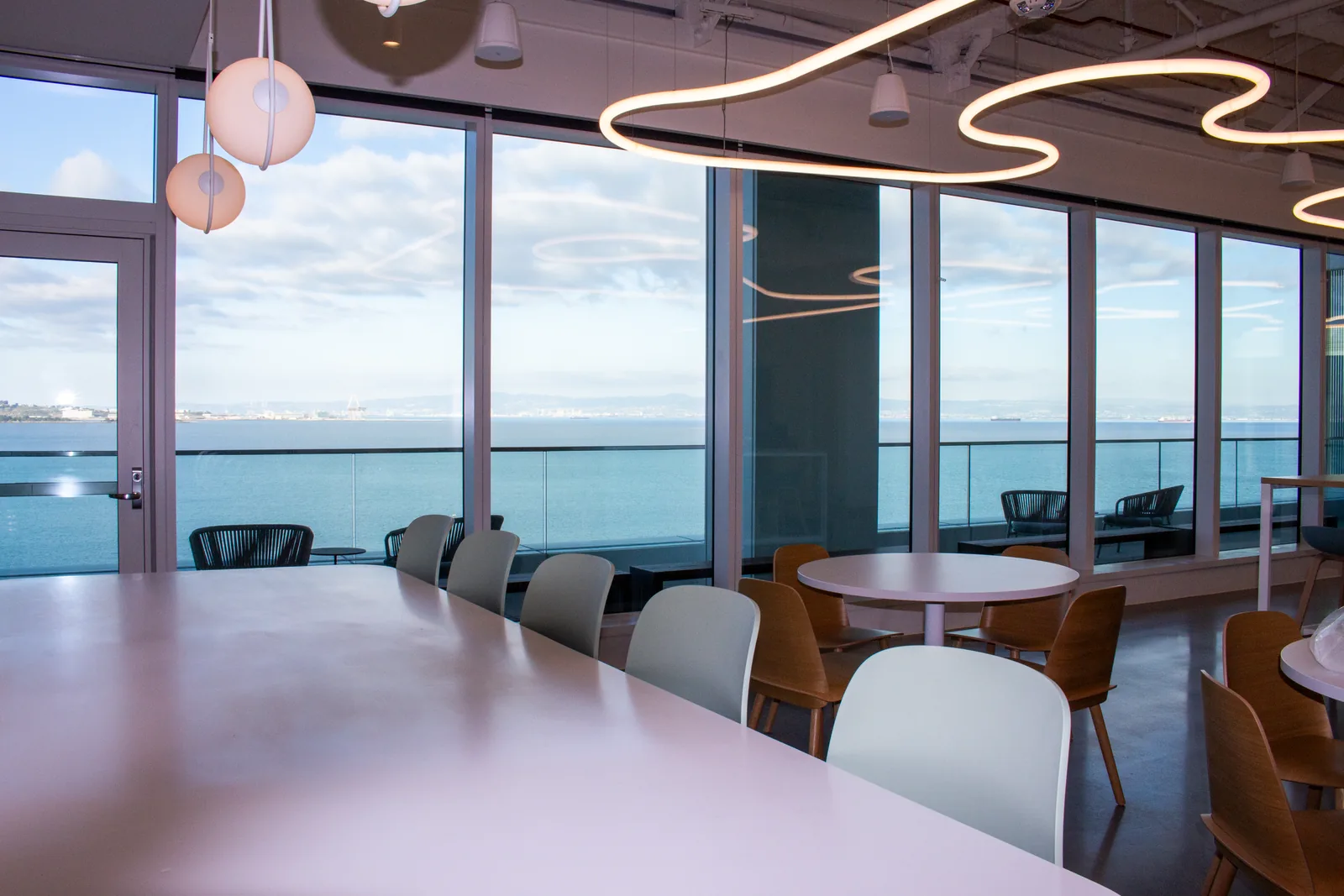 Modern San Francisco tech-office breakroom with floor-to-ceiling Bay views, sculptural pendant lighting, and a long communal dining table.